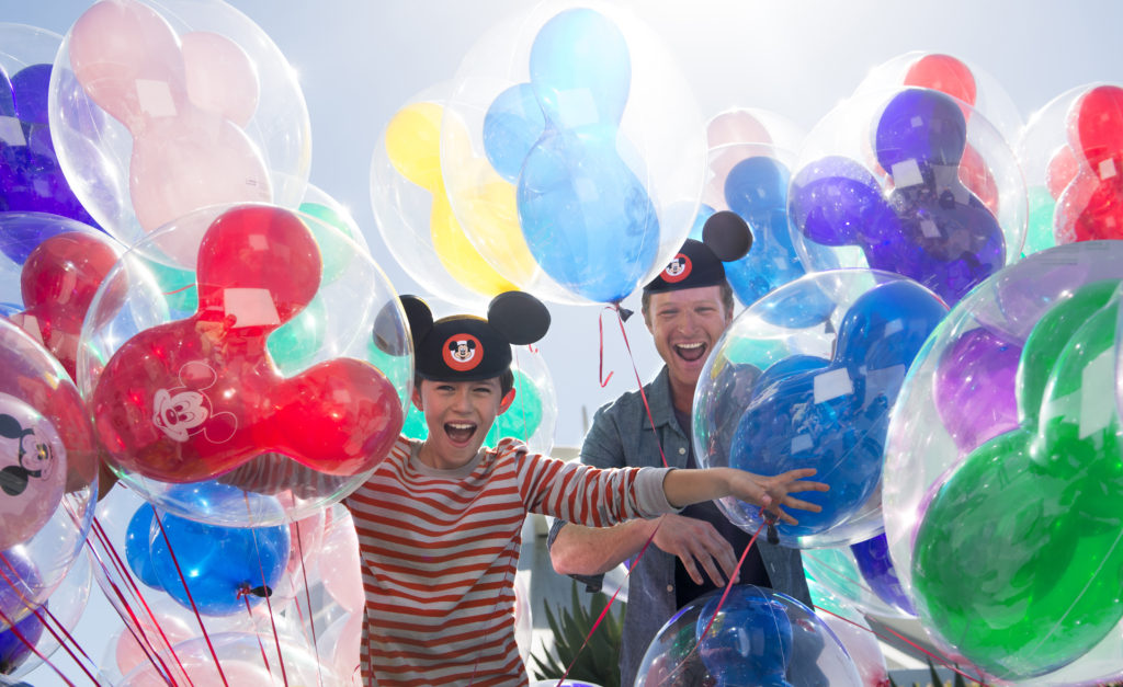 A Father and a Son both wearing Black Mickey Hats in a bunch of mickey balloons all different colors smiling and having fun